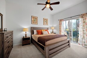 Une chambre à coucher avec un lit en bois, des tables de chevet, un ventilateur de plafond et une grande fenêtre avec des rideaux.