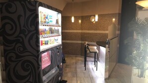 Una máquina expendedora con diferentes opciones de bocadillos y bebidas, un área de descanso con una silla y una mesa, y una planta en maceta.