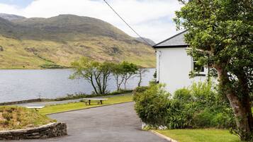 A white building with a stone wall and a paved path leading to a picnic table by a lake with mountains in the background.