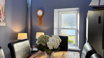 A dining area with a wooden table, chairs, a vase with flowers, a clock, and a window with a view of the outdoors.