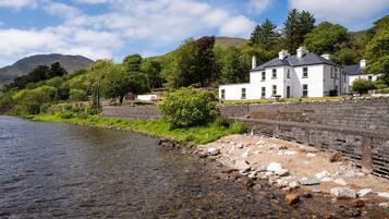 A riverside property with a stone wall, a white building with multiple windows, and a mountainous backdrop.