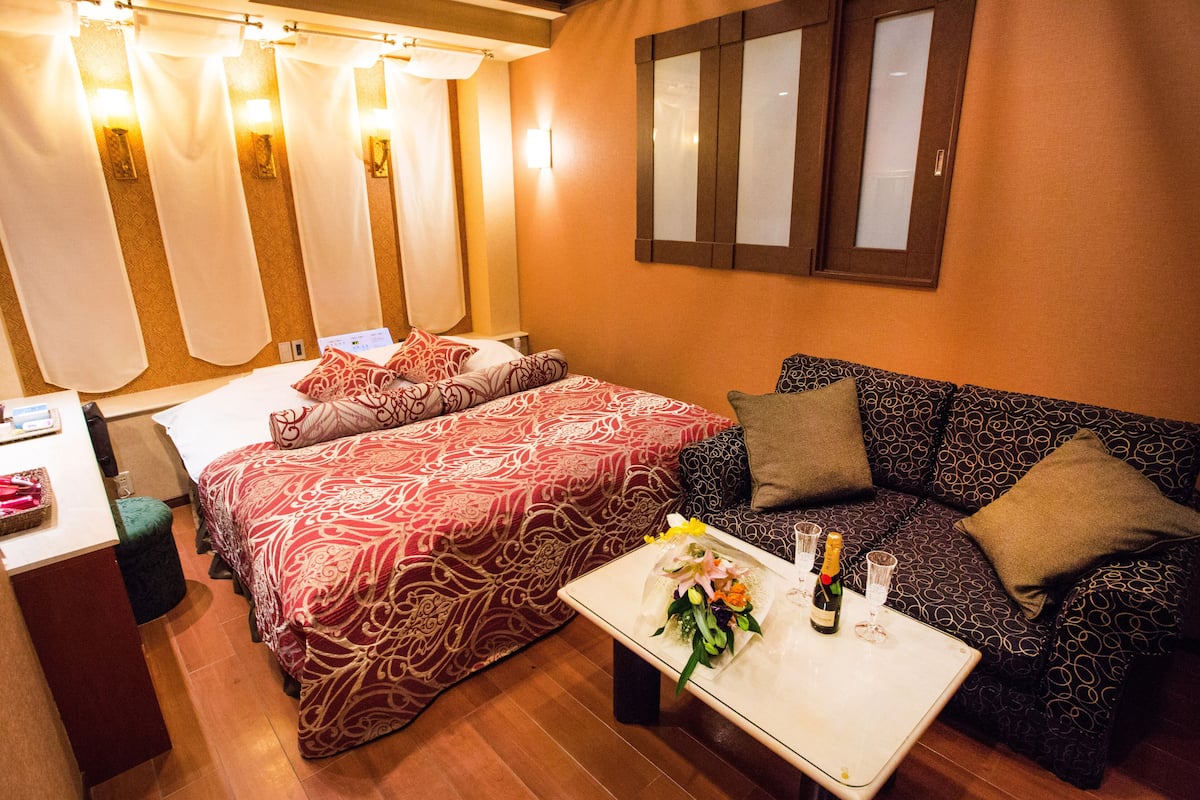 Ein Hotelzimmer mit einem großen Bett, einer Couch, einem kleinen Tisch mit Blumenstrauß und Champagner sowie einem Spiegel an der Wand.