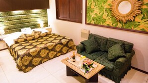 A hotel room with a bed, a green sofa, a coffee table with a bottle and glasses, and a wall with a decorative mirror and artwork.
