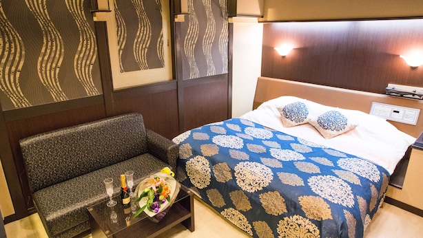 A hotel room with a patterned sofa, a bed with a blue and gold bedspread, and a small table with a bouquet of flowers and a bottle of champagne.