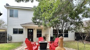 Un patio con sillas Adirondack rojas, un hogar de piedra y una casa al fondo.