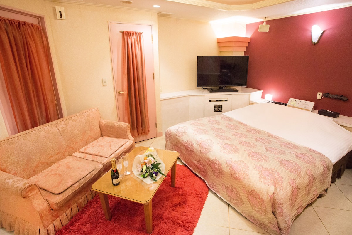 A hotel room with a sofa, a bed, a TV, and a small table with a bottle and flowers.