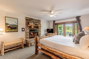 A bedroom with a stone fireplace, a bed with pillows, a television, and a view of the outdoors.
