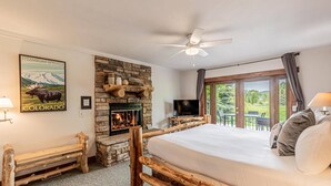 A bedroom with a stone fireplace, a bed, a television, and a balcony with a view of the outdoors.