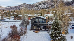 A modern lodge with a large deck and a mountain view, surrounded by snow-covered landscape and trees.
