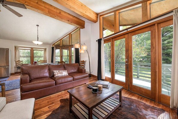 A spacious living room with a brown leather sofa, wooden coffee table, and large windows offering a view of the outdoors.