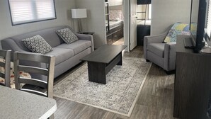 A modern living room with a grey sofa, a coffee table, and a TV. There is a window with blinds and a door leading to a bedroom. -  Niagara-on-the-Lake Cottage (Niagara-on-the-Lake)