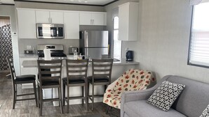 A modern kitchen with white cabinets, a stainless steel refrigerator, and a microwave. There is a dining area with a wooden table and chairs. A gray sofa with patterned pillows is in the living area. -  Niagara-on-the-Lake Cottage (Niagara-on-the-Lake)