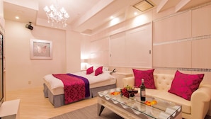 A modern hotel room with a glass coffee table, a bed with a pink and purple blanket, a white sofa with pink pillows, and a chandelier. - Restay Koriyama - Adults Only (Koriyama)