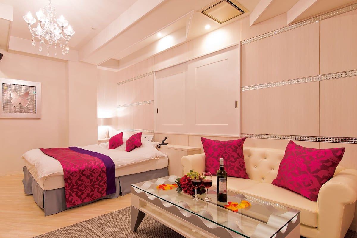 A modern hotel room with a glass coffee table, a bed with a pink and purple blanket, a white sofa with pink pillows, and a chandelier.