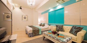 A modern hotel room with a glass coffee table, a sofa, a bed with a patterned bedspread, and a chandelier.