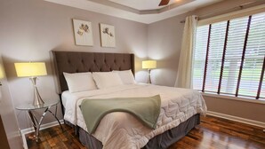Ein ordentlich eingerichtetes Schlafzimmer mit einem großen Bett, zwei Nachttischlampen, einem Fenster mit Jalousien und zwei an der Wand hängenden Blumenbildern.