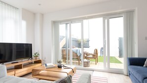 Una sala de estar moderna con una televisión de pantalla plana, muebles de madera, un sofá azul y un puerta corrediza de vidrio que da a un patio con mobiliario exterior.