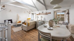 A cozy living room with a fireplace, a dining table, and a high ceiling with exposed beams. - Carvossa - Sea Views & Woodburner (Marazion)