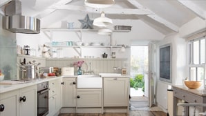 A kitchen with a central island, white cabinetry, and a rustic wooden floor. There is a window providing natural light and a view of greenery outside. - Carvossa - Sea Views & Woodburner (Marazion)