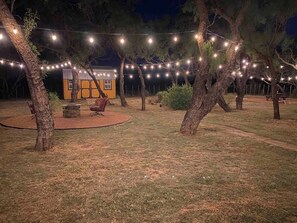 Un espacio al aire libre con luces colgantes, un edificio pequeño y un brasero.