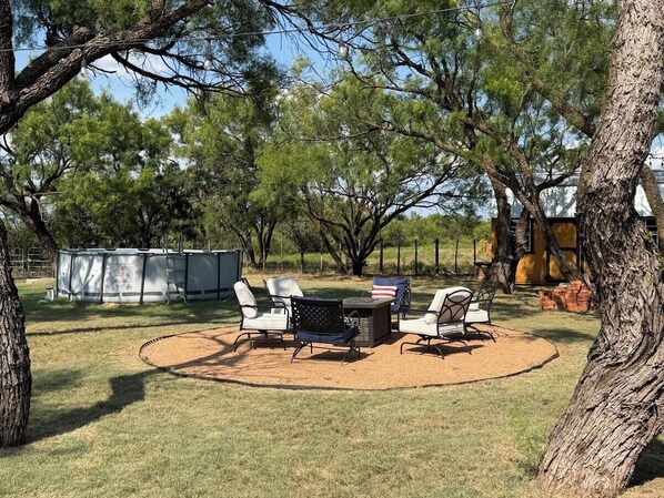 An outdoor patio area with a round table and chairs, surrounded by trees and a fenced yard.