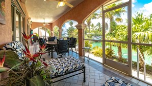 Un patio luminoso con mesa y sillas, rodeado de amplios ventanales que ofrecen vistas a una piscina y palmeras.