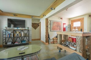 A living room with a round glass coffee table, a bookshelf, a flat-screen TV, and a kitchen area with a wooden countertop and various kitchen items.