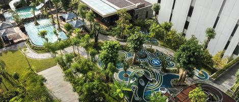 An aerial view of a resort with multiple swimming pools, a water slide, and a playground area surrounded by lush greenery.