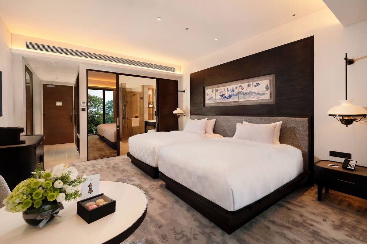 A modern hotel room with two beds, a desk, a chair, a small table with a vase of flowers, and a painting on the wall.