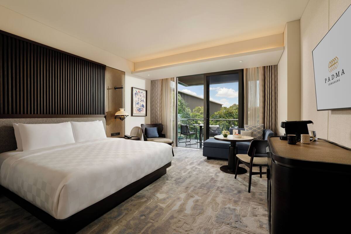 A hotel room with a large bed, a desk, a chair, a small table, and a balcony with a view.