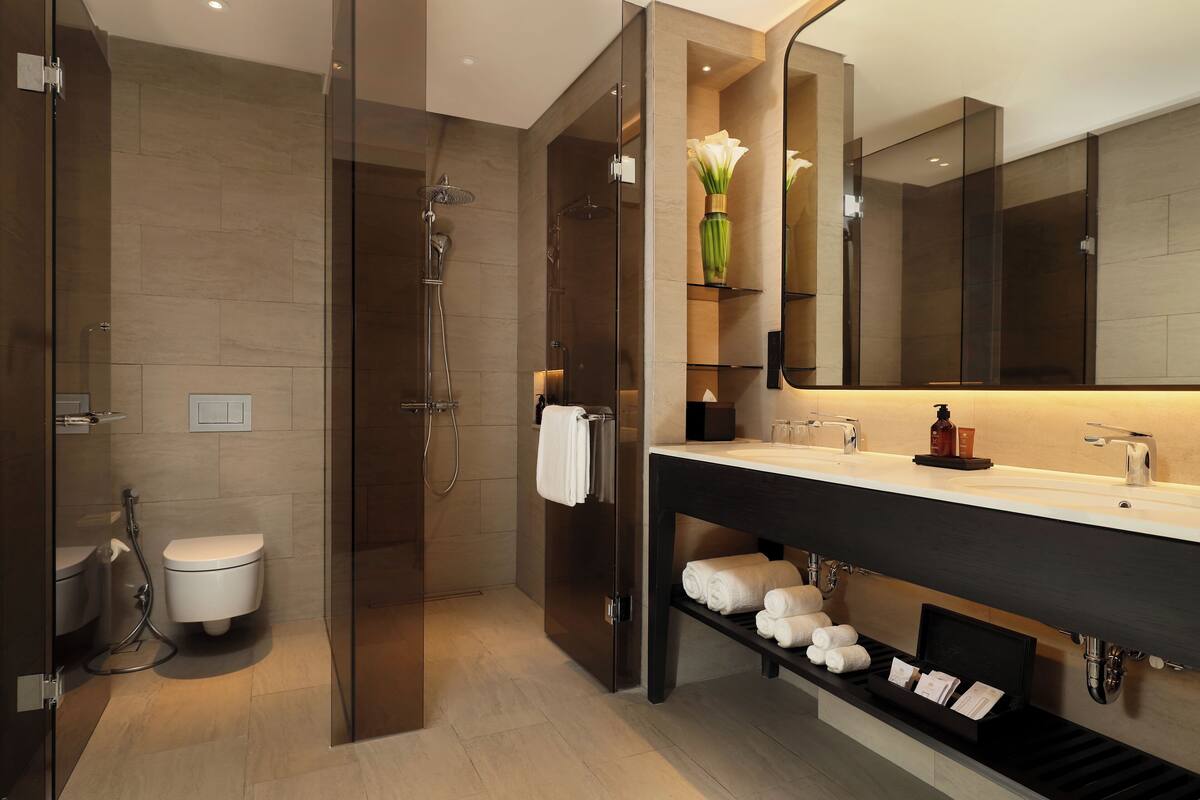 A modern bathroom with a glass-enclosed shower, a white toilet, a vanity with a large mirror, and a shelf with neatly folded towels.