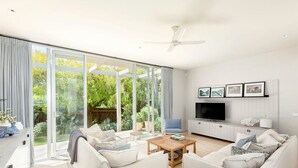Amplio salón con un gran ventanal corredero que da a un jardín, un televisor de pantalla plana y un ventilador de techo.