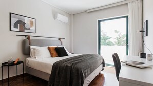 A modern bedroom with a bed, bedside table, and a desk with a chair. There is a window with curtains, a wall-mounted air conditioner, and a framed picture on the wall. - Top-Class 2BR Apartment in Marousi (Marousi)