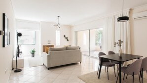 A modern living room with a sofa, dining table, and chairs. There is a large window with curtains, a floor lamp, and a ceiling light. - Top-Class 2BR Apartment in Marousi (Marousi)