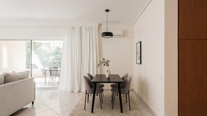 A modern living room with a sofa, dining table, and chairs. There is a large window with curtains, a hanging light fixture, and a wall-mounted air conditioner. - Top-Class 2BR Apartment in Marousi (Marousi)