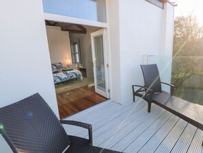 A balcony with two wicker chairs, a wooden deck, and a bedroom with a bed and nightstand.
