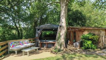 A covered outdoor seating area with a bench and table, surrounded by trees and a wooden structure.