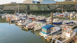 A marina with multiple boats docked, a large industrial building in the background, and a stone wall along the water's edge.