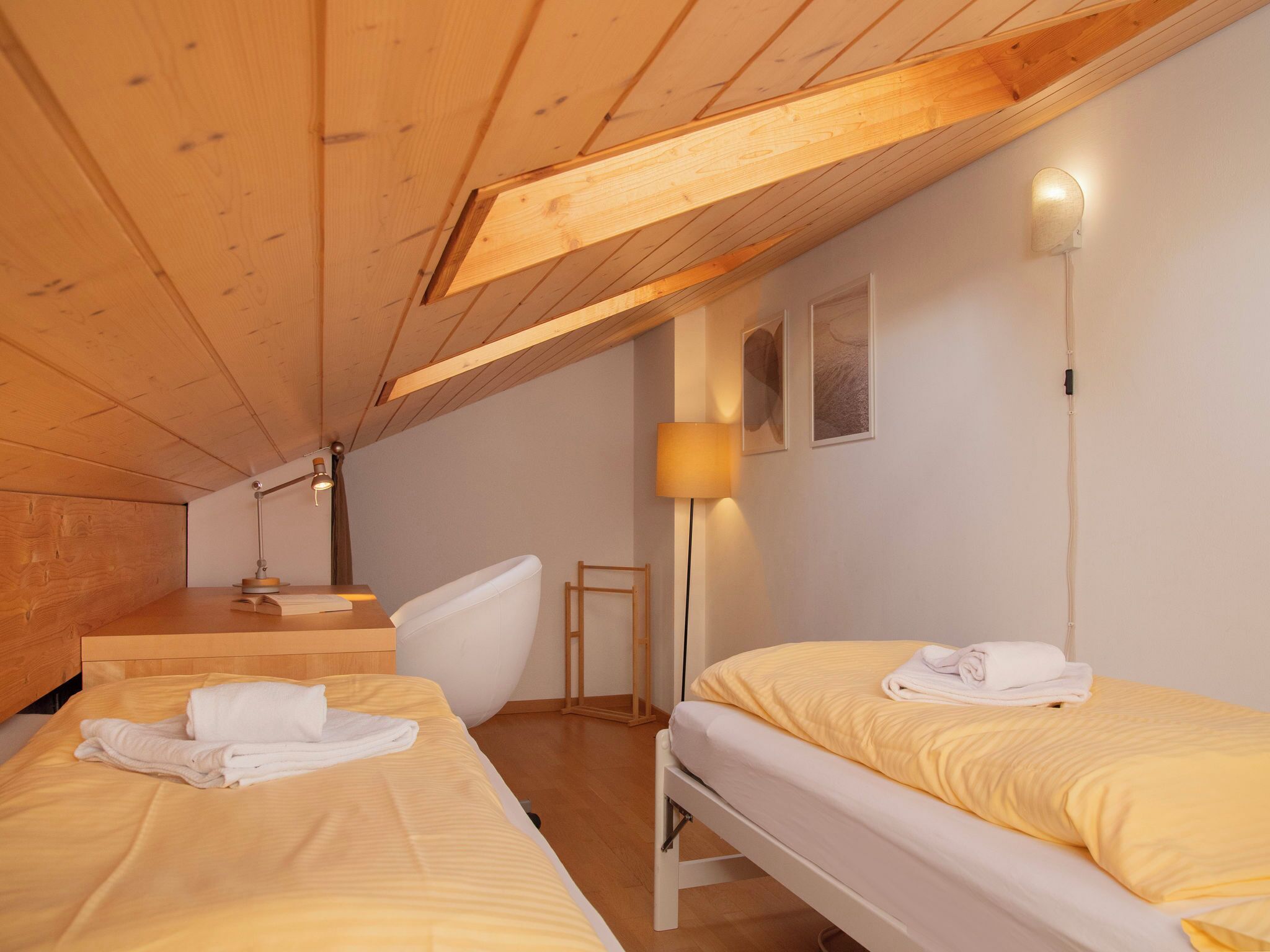A cozy room with wooden beams, two beds with yellow bedding, a white chair, a desk, and a lamp.