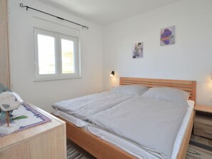 Una cama individual con cabecera de madera en una habitación con paredes blancas. Hay una ventana con persianas blancas, una mesita con un objeto decorativo y dos lámparas fijadas a la pared.