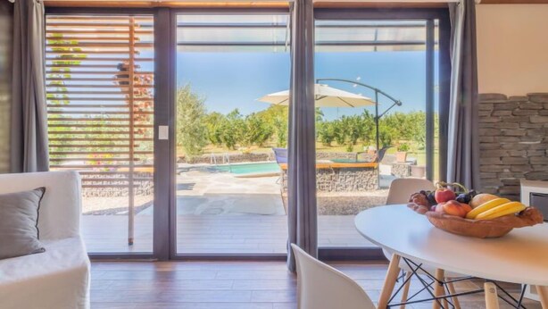 A room with a sliding glass door leading to an outdoor pool area, a dining table with a fruit bowl, and a stone wall.