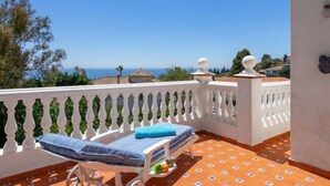 A balcony with a white balustrade, a lounge chair with a striped cushion, and a view of the sea and distant buildings. - Villa Juna by Interhome (Benalmádena Costa)