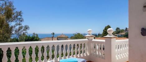 A balcony with a white balustrade, a lounge chair with a striped cushion, and a view of the sea and distant buildings.