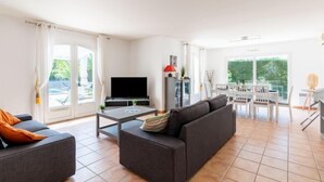 A modern living room with a sofa, a coffee table, and a flat-screen TV. There is a dining area with a table and chairs, and a balcony with a view of greenery. - Massenet by Interhome (Saint Aygulf)