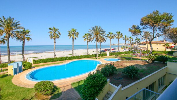 A swimming pool surrounded by palm trees, a beach, and a building with a balcony. - Beautiful apartment in First Line Marina Azul (Els Poblets)