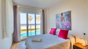 A hotel room with a bed, a painting, a nightstand, and a view of a pool and beach. - Beautiful apartment in First Line Marina Azul (Els Poblets)