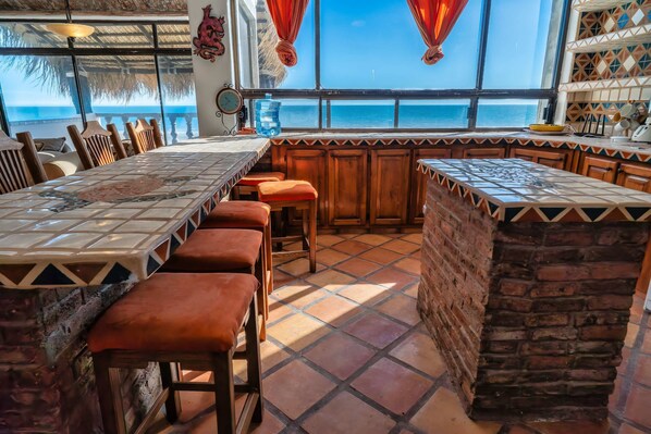 A restaurant with a long dining table, cushioned bar stools, and a view of the ocean.