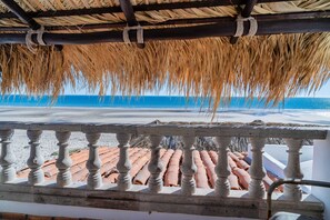 A balcony with a thatched roof overlooking a beach with terracotta tiles.