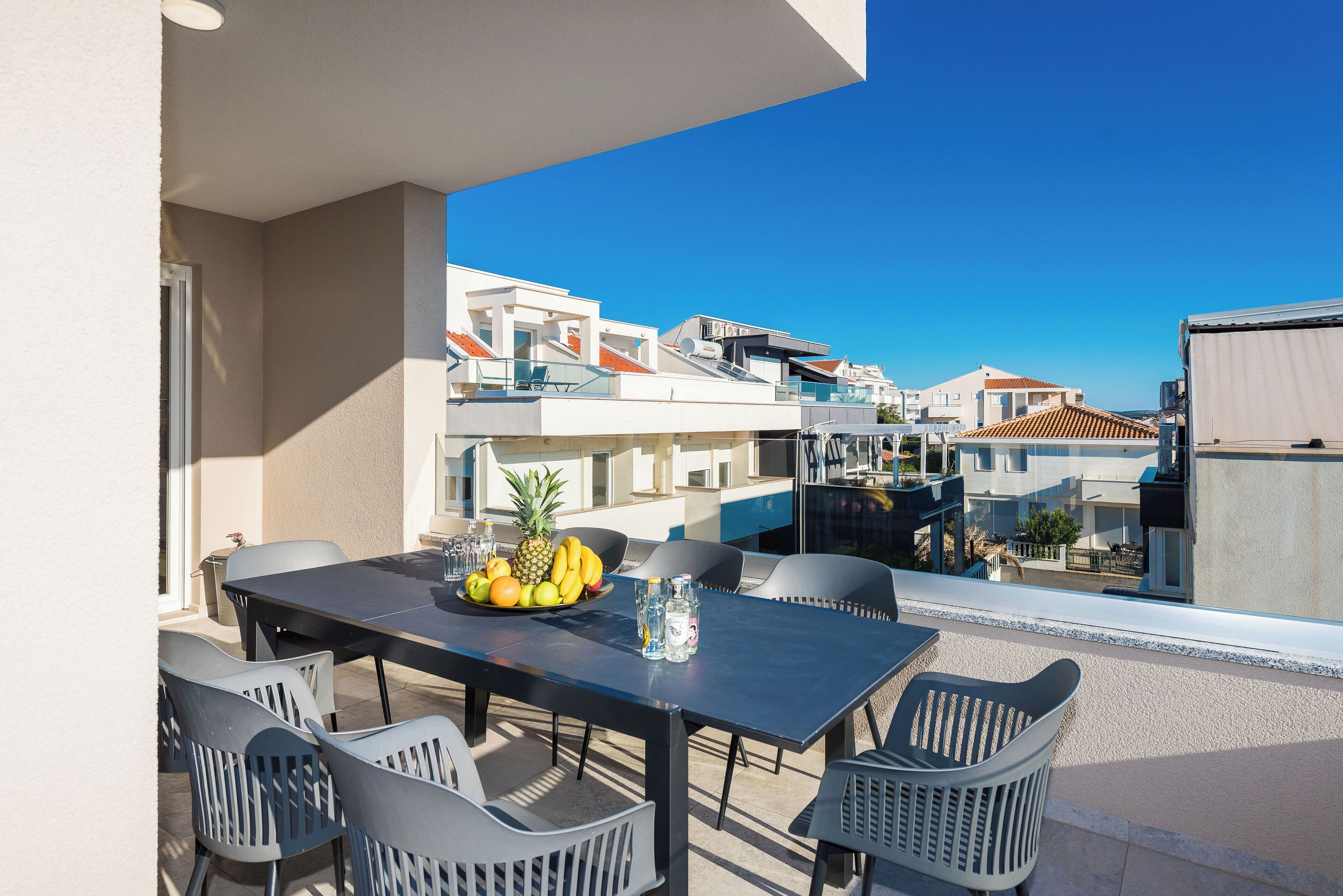 An outdoor balcony with a dining table set for four, surrounded by chairs, and a fruit bowl on the table.