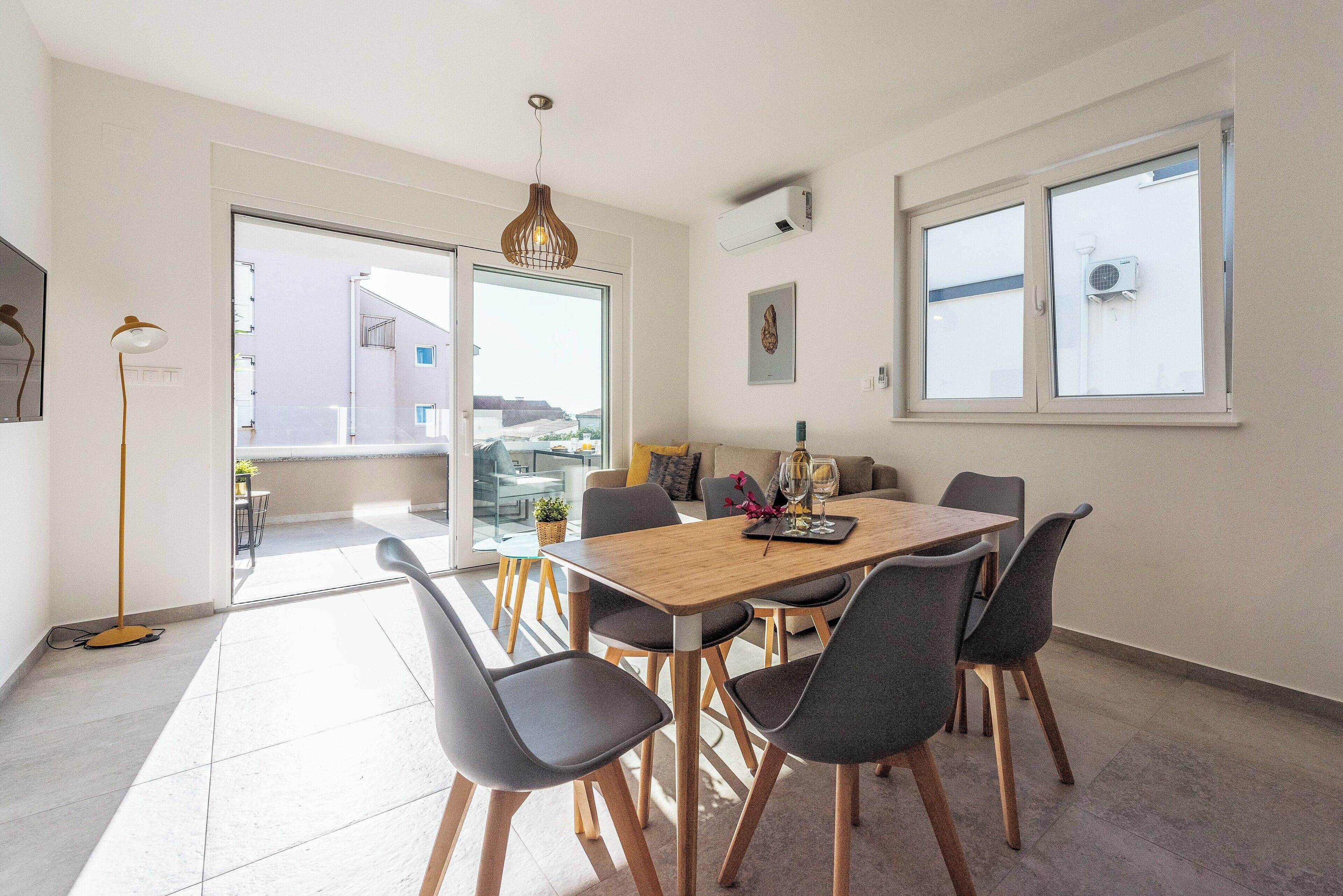 A modern dining area with a wooden table and grey chairs, large windows, and an air conditioning unit on the wall.